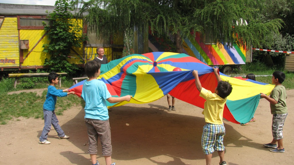 Kinder und ein Pädagoge spielen mit einem bunten Riesentuch. Im Hintergrund Bauwägen auf dem Abenteuerspielplatz.